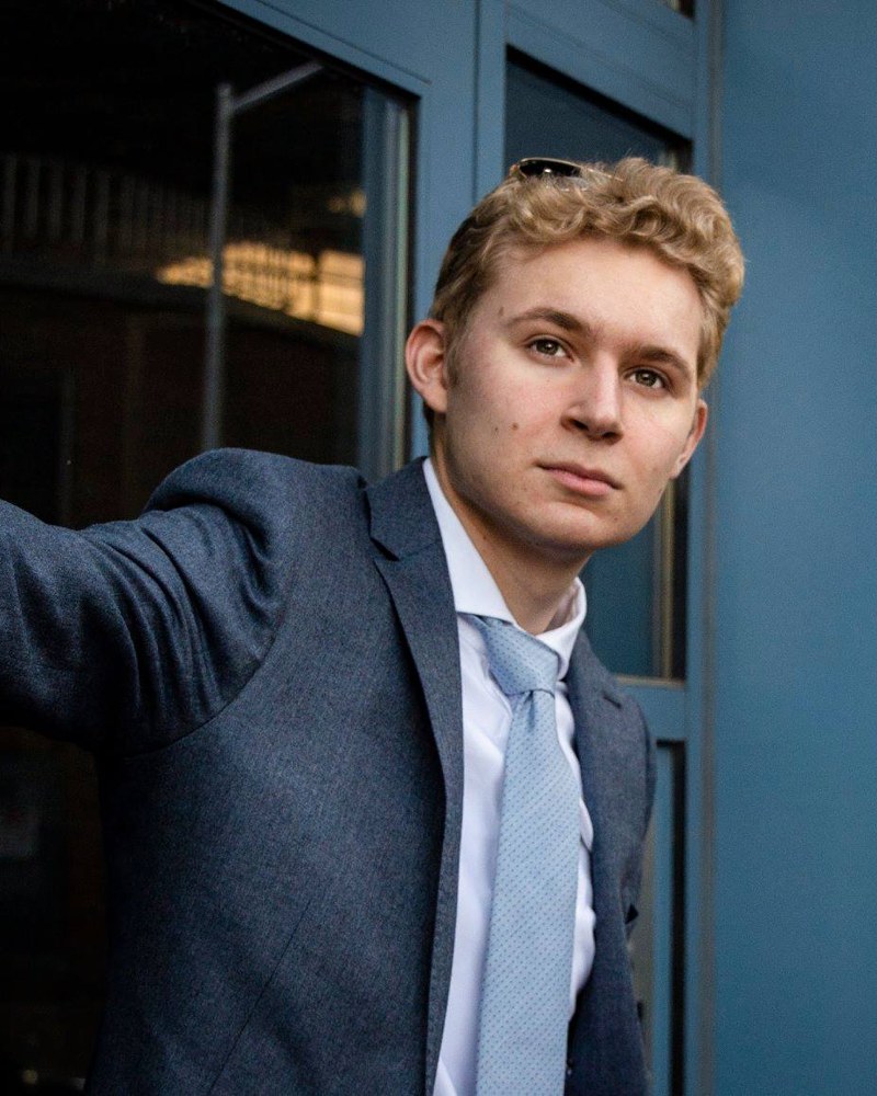 Meric in a navy blazer and light blue tie, leaning out from a blue doorway